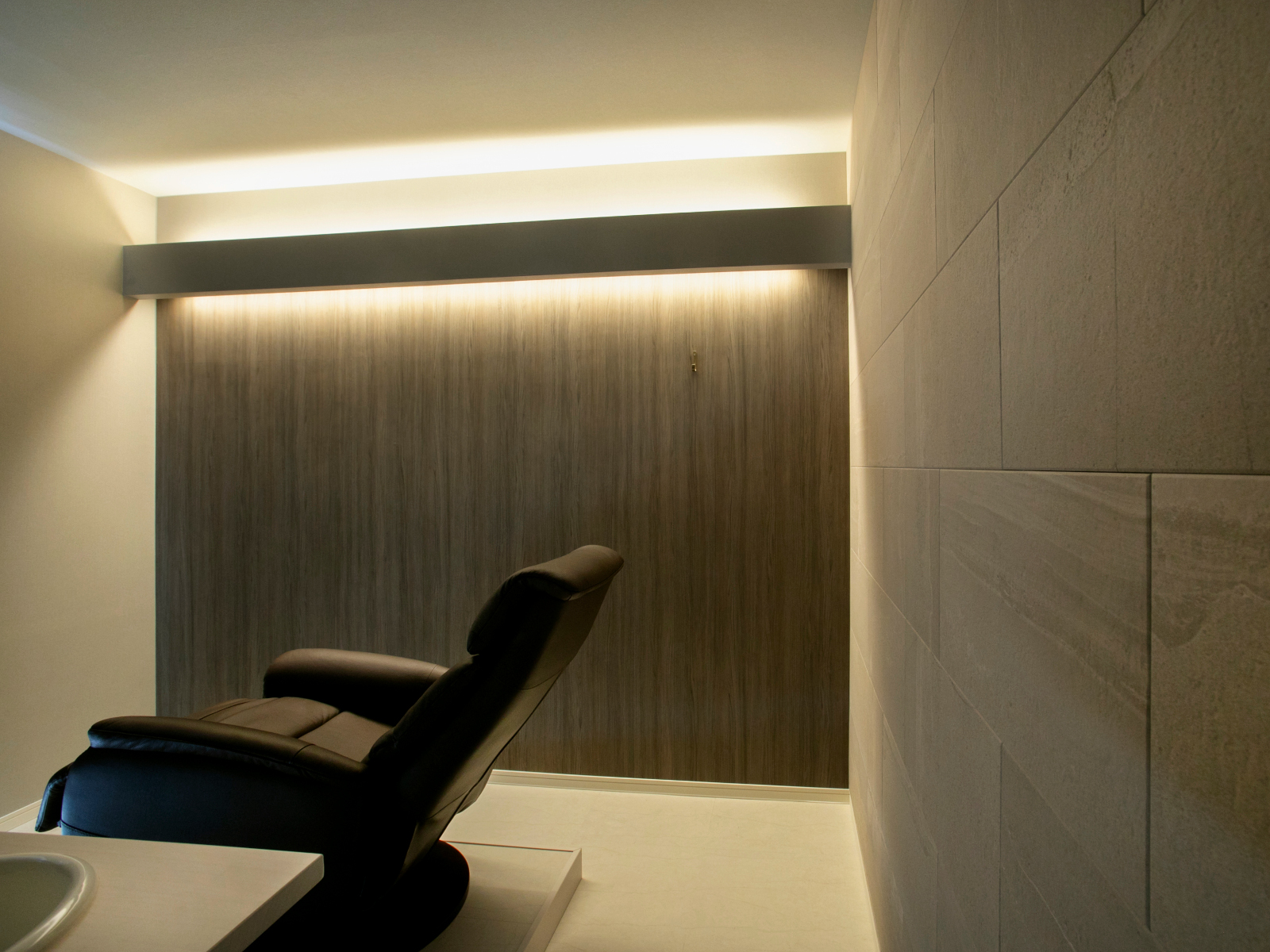 ネイルサロンリフォーム。エコカラットストーングレース ベージュと木目調壁を組み合わせ、間接照明が柔らかく照らす落ち着いたサロン空間。