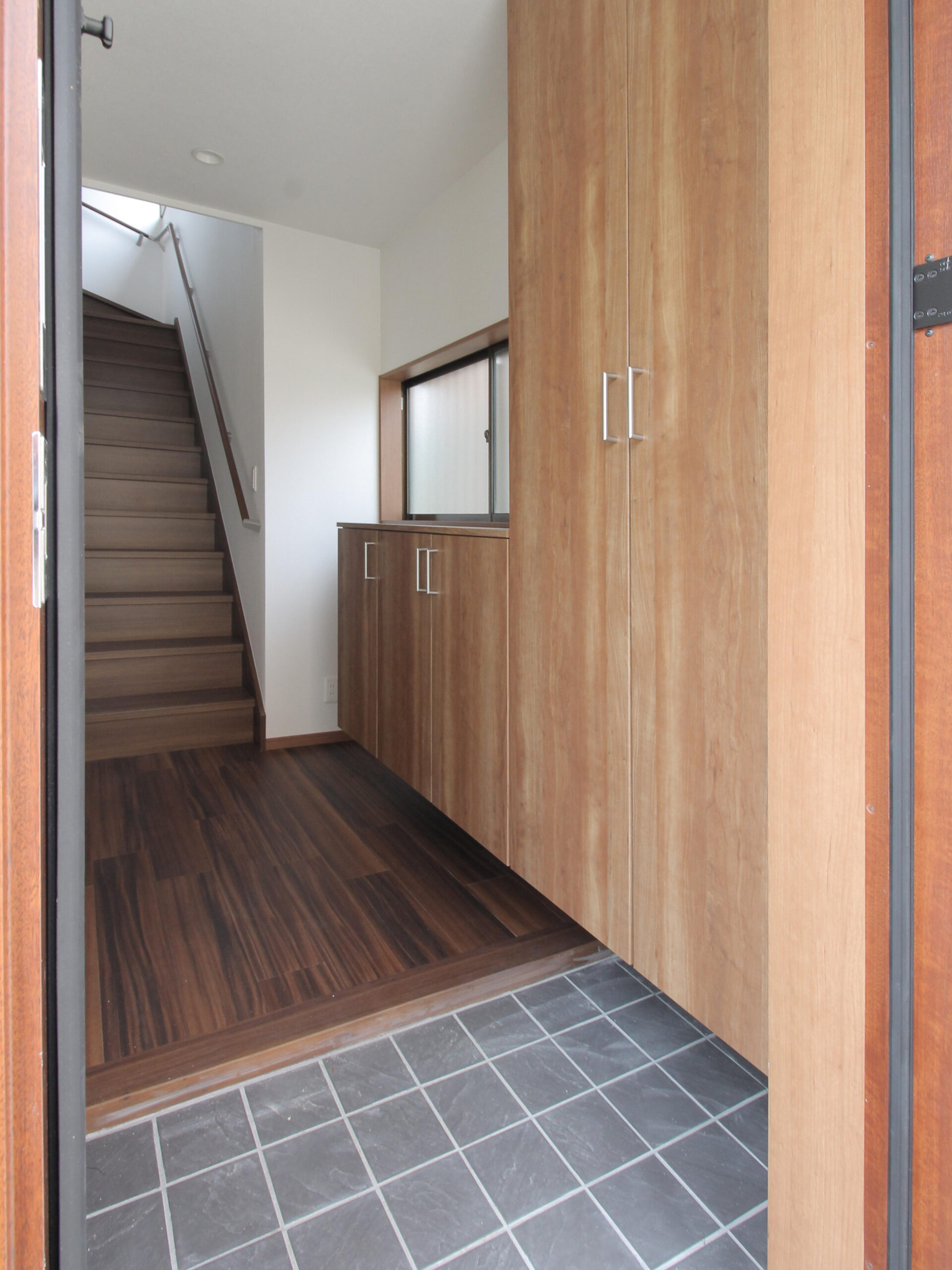 玄関土間タイルと木目の玄関収納が調和した玄関ホール。戸建ての階段をゆるやかにしたリノベーションの施工事例。